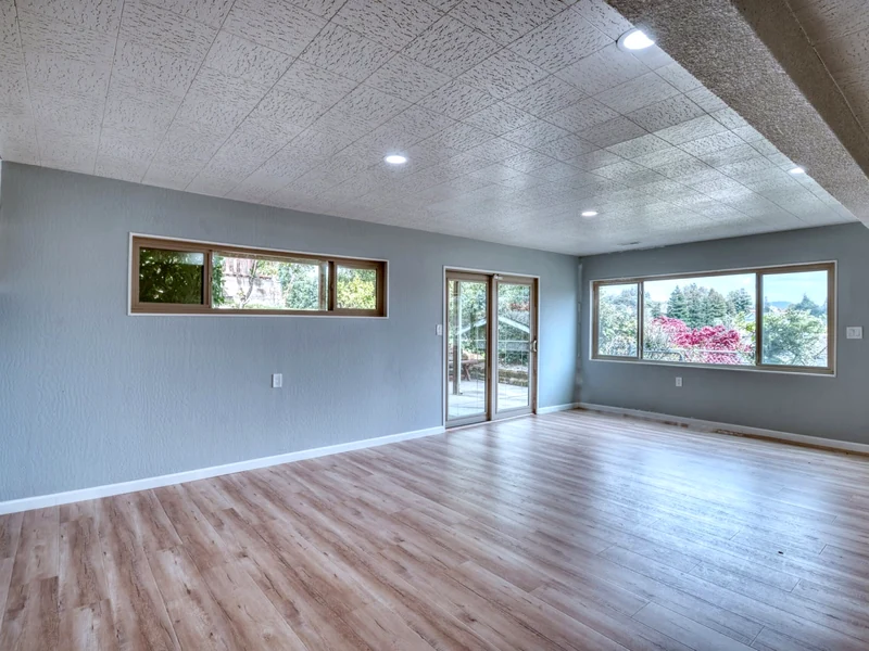 Spacious empty room with gray walls, light wood flooring, recessed ceiling lights, large windows, and sliding glass doors.