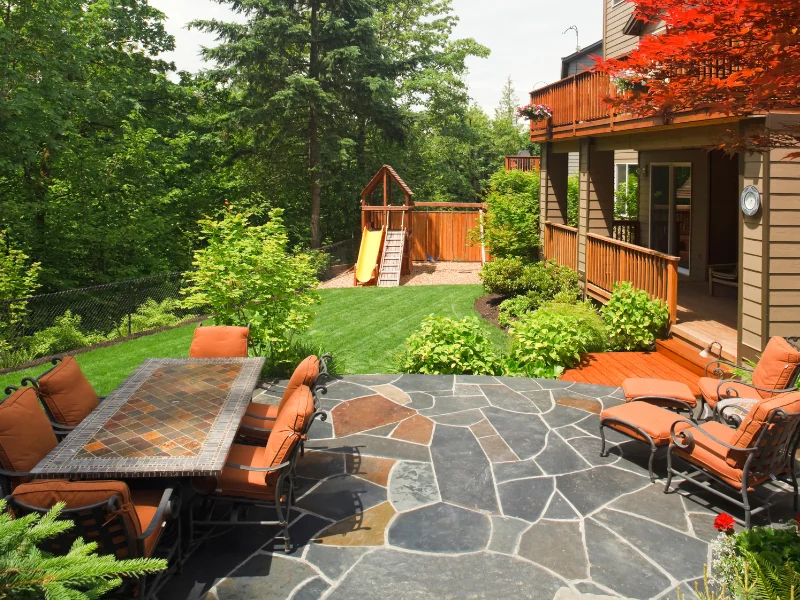 Backyard flagstone patio with a tile-top dining table and orange cushioned chairs, a wooden deck, and a kids’ playset on the lawn.