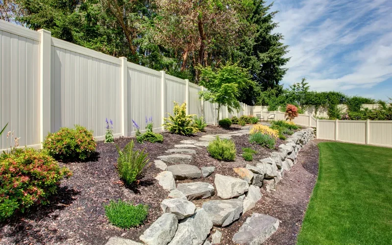 Landscaped backyard with a stepping-stone path through mulched flower beds, rock edging, and a cream privacy fence beside a green lawn.