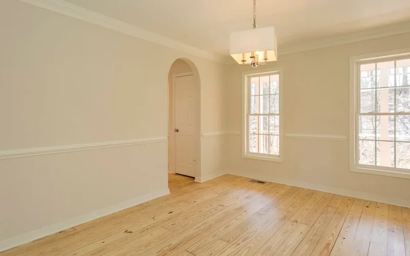 Empty dining room with light hardwood flooring, neutral walls, two large windows, crown molding, and a modern pendant light.