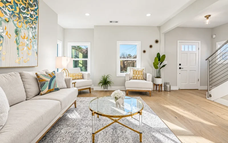 Bright modern living room with neutral seating, glass coffee table, light wood flooring, colorful wall art, and a contemporary staircase.