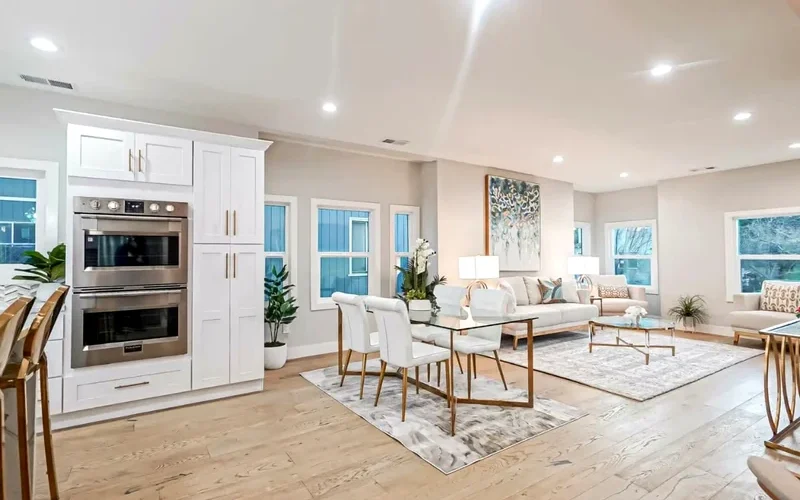 Open-concept living and dining area with light wood floors, white cabinetry, neutral furniture, recessed lighting, and large windows.