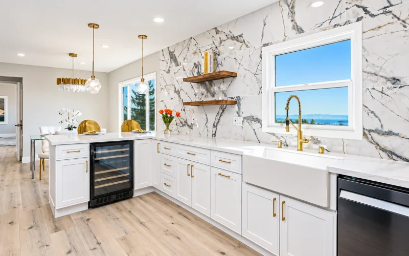 Bright modern kitchen with white shaker cabinets, marble-look backsplash, farmhouse sink, gold fixtures, and light wood flooring.