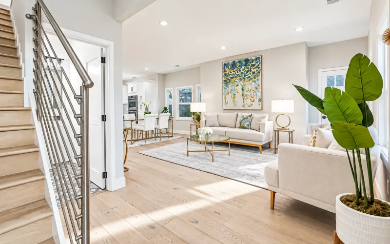Open-concept living room with light wood floors, neutral furniture, modern metal staircase railing, large indoor plant, and adjacent dining area.