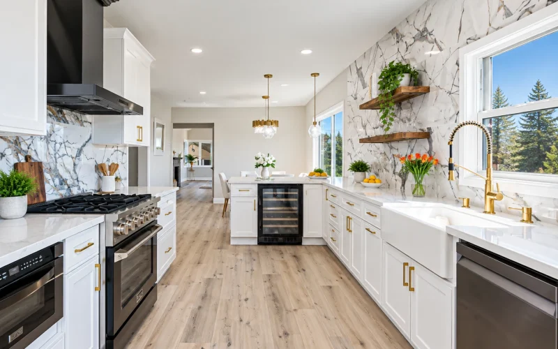 Spacious modern kitchen with white shaker cabinets, marble-look backsplash, farmhouse sink, gold fixtures, stainless steel appliances, and light wood flooring.