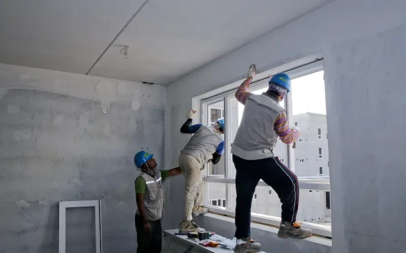 Three construction workers in hard hats positioning and securing a new window in an unfinished interior wall during window installation.