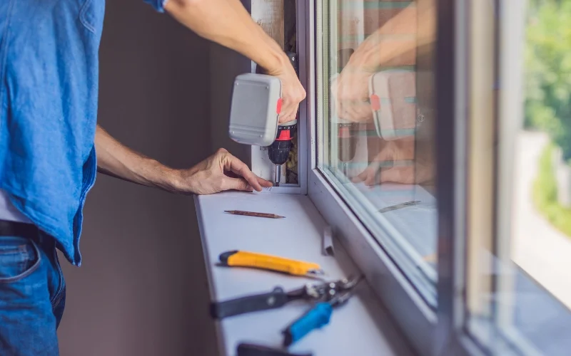 Technician using a cordless drill to secure a window frame during window repair and installation, with a utility knife, pliers, and screwdriver laid out on the sill.